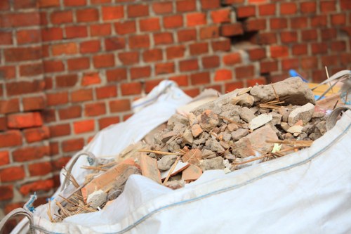 A large, white, heavy-duty sack filled with a pile of construction debris, including broken bricks, concrete fragments, and small wooden sticks, resting on a flat surface outdoors. The debris is varied in size and color, predominantly light beige and reddish-brown, with some dust and dirt visible on the materials. The sack is situated against a background of red brick wall with a textured surface, indicative of a building exterior or driveway area, possibly within a residential or commercial property in South Kensington. The lighting is natural, highlighting the rough textures of the rubble and the woven fabric of the sack. This scene reflects the type of waste commonly collected during house clearance or renovation projects, consistent with rubbish removal services offered by South Kensington House Clearance.