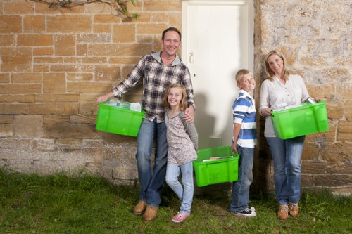 A family of four standing outdoors against a brick wall, each holding a bright green plastic storage container used for rubbish collection or clearance. The father is on the left, smiling and wearing a checkered shirt and jeans, with a container in his right hand. Next to him is a young girl with long hair, dressed in a grey sweater and light blue jeans, holding a container in her left hand. To her right is a young boy wearing a blue and white striped shirt and dark trousers, clutching a container close to his chest. On the far right is a woman with blonde hair in a white, patterned blouse and jeans, also holding a green rubbish container. The background features a stone and brick exterior wall with a white wooden door in the center, and the family is standing on a grassy patch. The scene reflects a demonstration of rubbish removal or house clearance, aligning with services offered by South Kensington House Clearance in the local area near South Kensington, London, with the environment being natural daylight and a casual, residential setting.