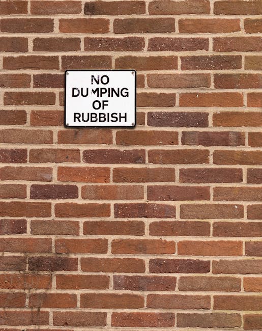 A large, overflowing collection of mixed household waste and recycling bags, cardboard boxes, paper, and various discarded items piled in a public outdoor space near a parking area. The rubbish includ