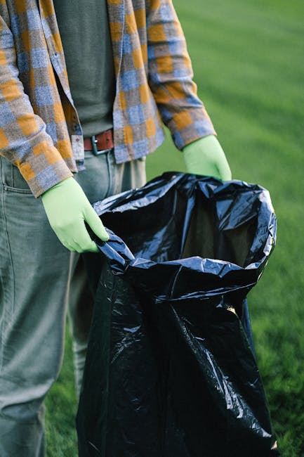 A person wearing a plaid shirt in shades of grey, yellow, and blue, paired with green gloves, is holding open a large black garbage bag outdoors on a grassy area. The individual is wearing grey pants 